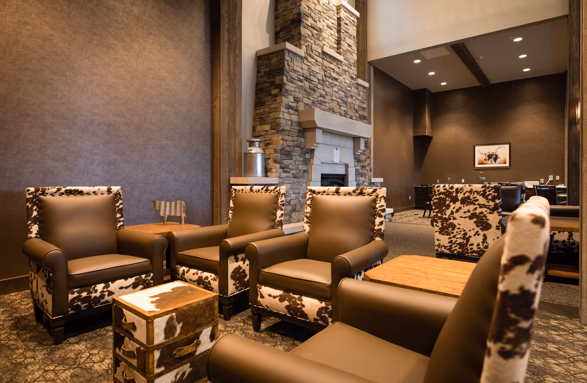 A cozy senior living facility lounge area with brown leather and cowhide-patterned armchairs arranged around a small wooden table. The room features a stone fireplace and warm lighting, creating a welcoming atmosphere.