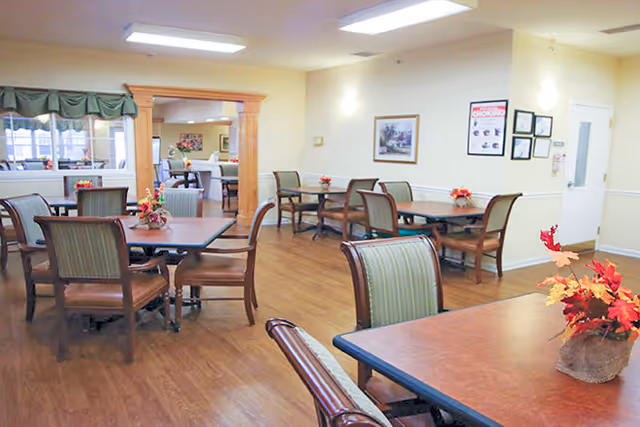 Dining room in a senior living facility with multiple tables and chairs, hardwood floors, and floral centerpieces.