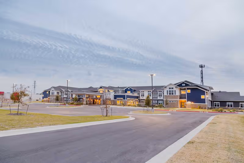 Exterior view of a large, modern senior living facility building with multiple windows and a covered entrance, surrounded by a paved driveway and landscaped grass areas under a cloudy sky.