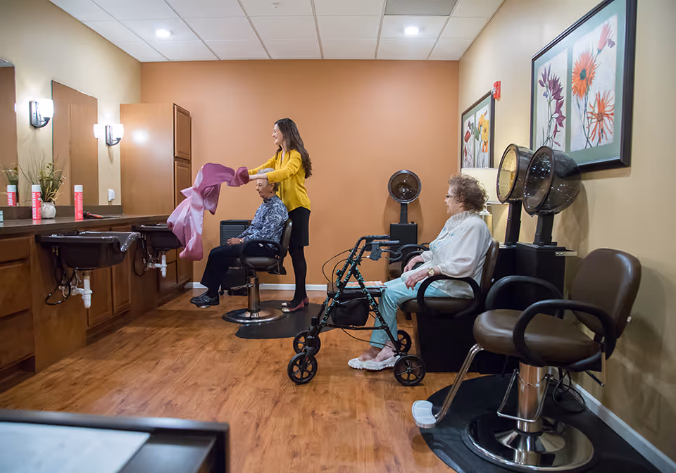 A senior living facility hair salon where a woman in a yellow top is placing a cape on an elderly man seated in a salon chair. Another elderly woman with a walker is seated nearby under hair dryers. The room has wooden floors, beige and orange walls, and framed floral artwork on the wall.