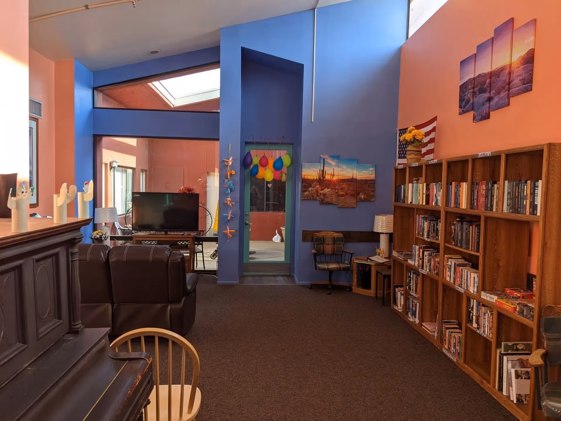 A cozy living room area with blue and orange walls, featuring a bookshelf filled with books, a plaid chair, a TV on a stand, and a piano with a wooden chair. Colorful balloons and decorations hang on a blue wall near a glass door leading to an outdoor courtyard.