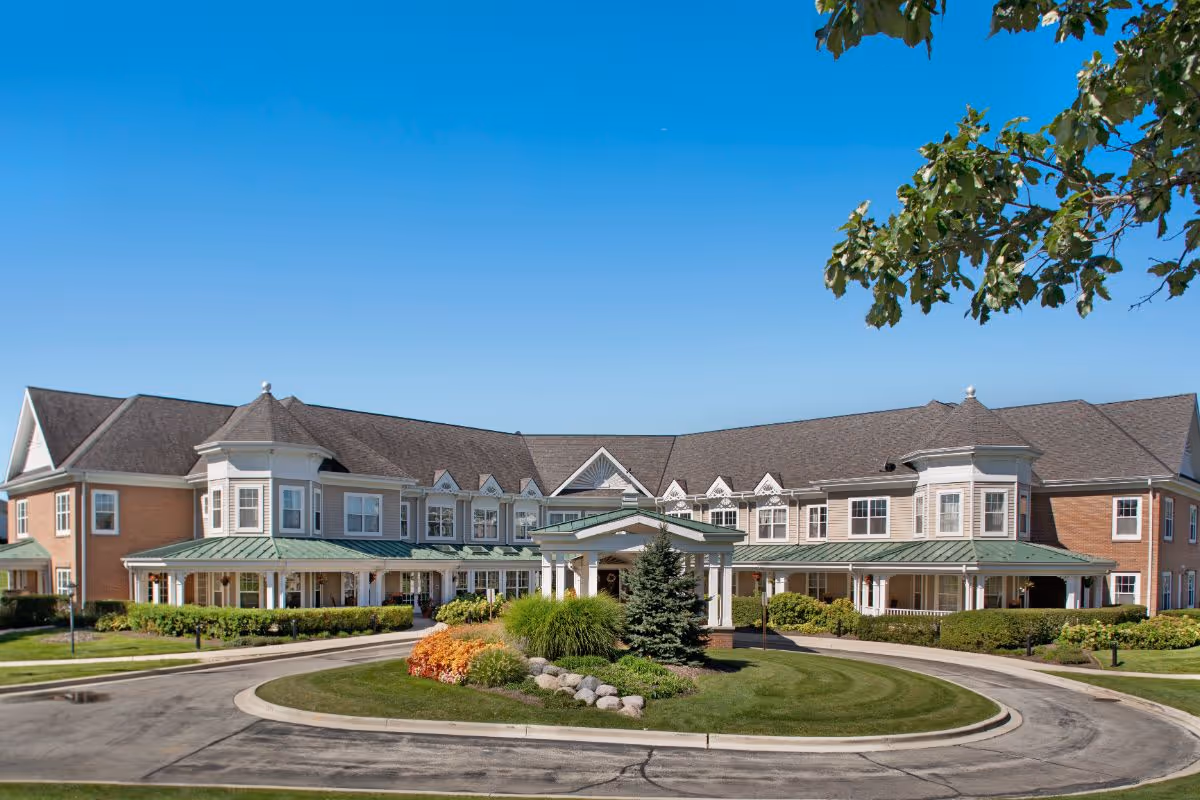 Front exterior of a large two-story senior living building with a covered entrance, circular driveway, and landscaped grounds.