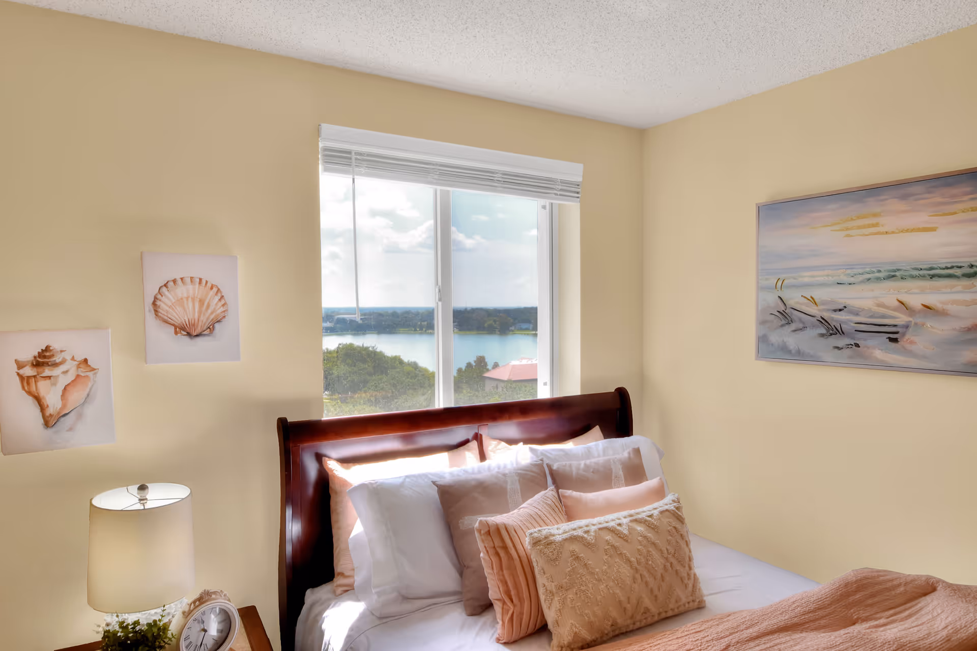 Sunlit bedroom with a wooden sleigh bed, layered pillows and throw, a bedside table with lamp and clock, coastal wall art, and a window showing a lake view.