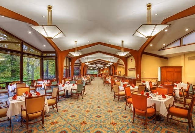 Large formal dining room with rows of set tables and chairs, patterned carpet, vaulted ceiling with wooden beams, and tall windows looking out to trees.