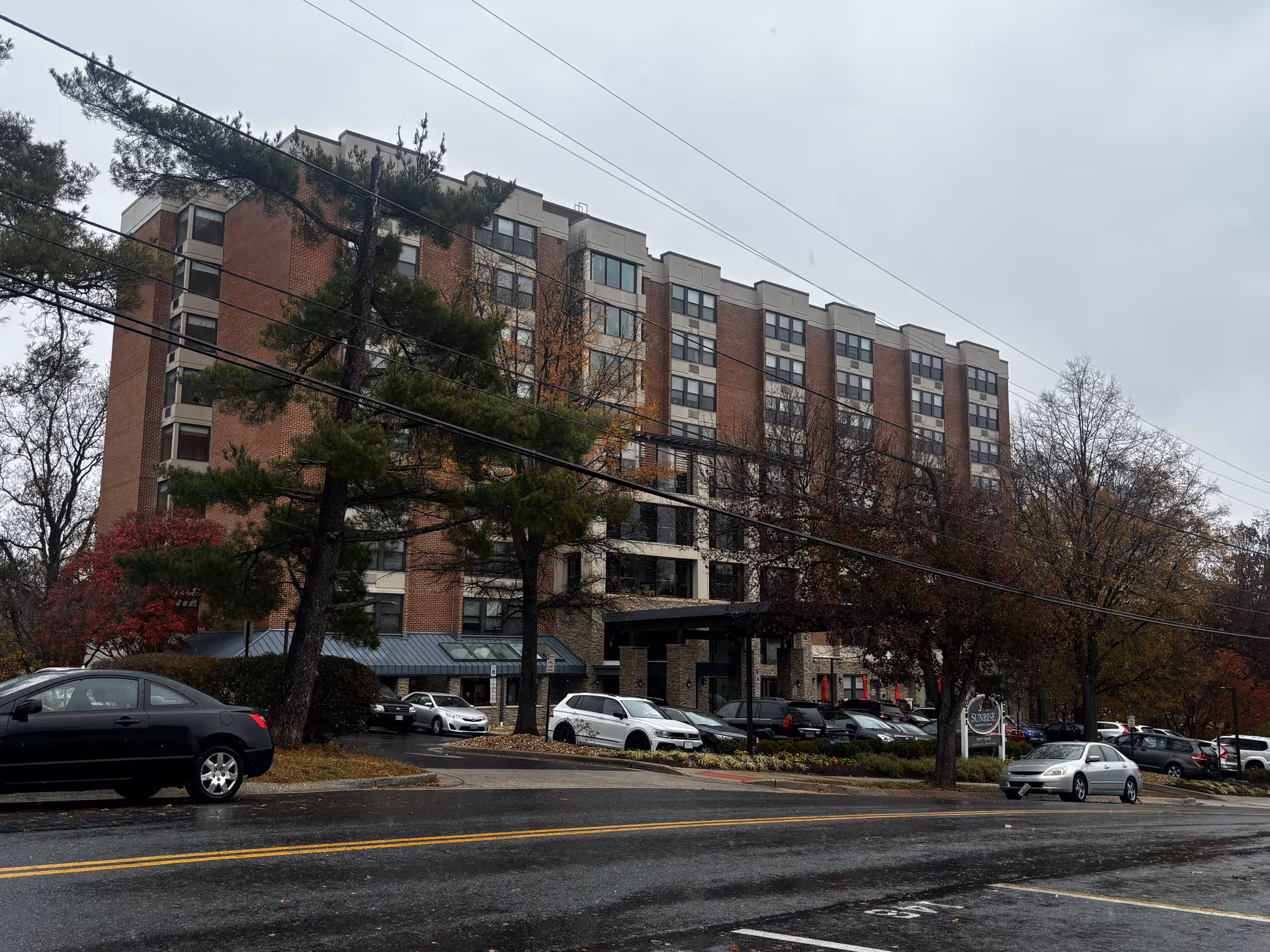 Street view of a multi-story brick residential building with parked cars, trees, and a wet road on an overcast day.