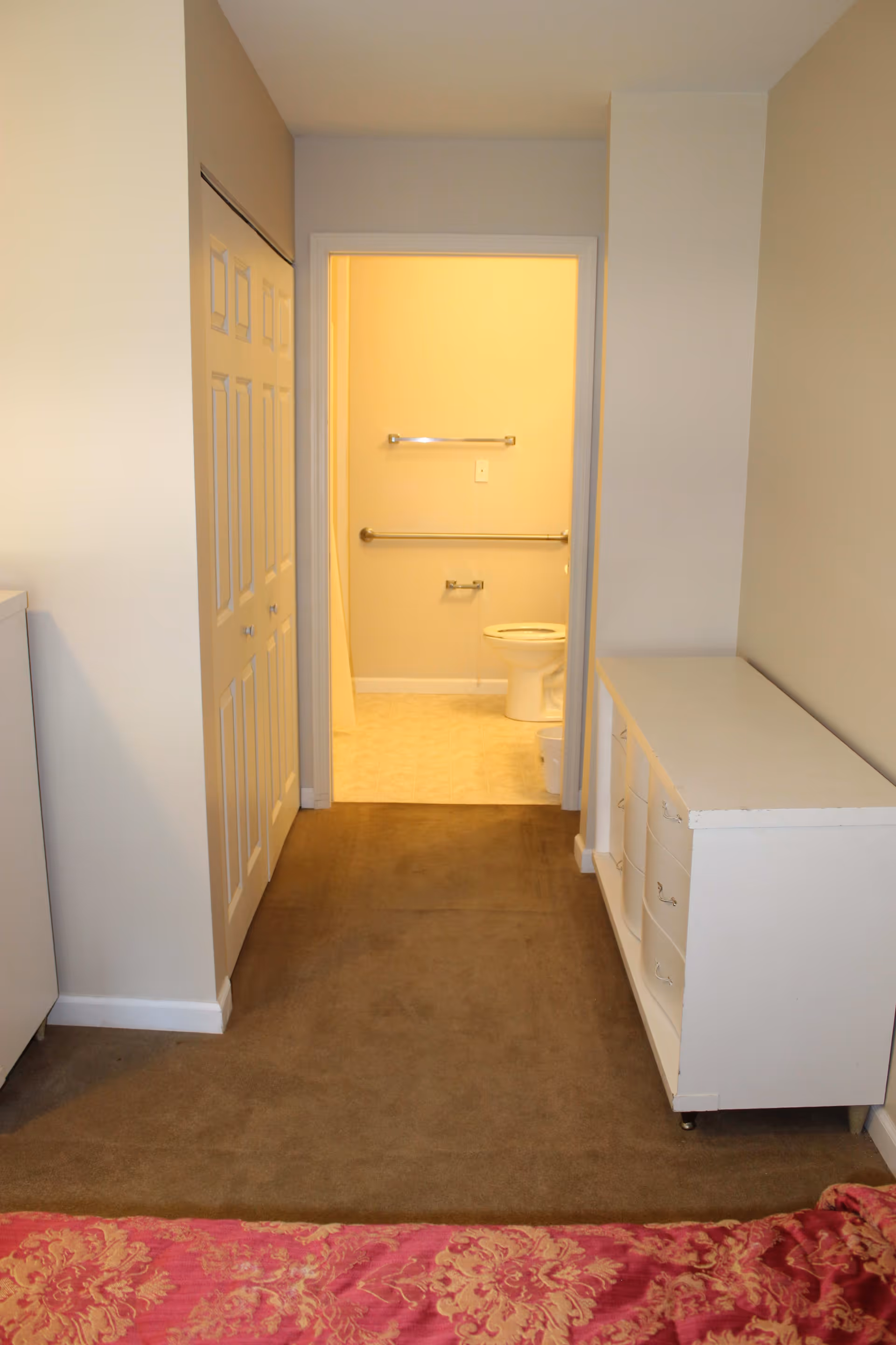 View from a bedroom looking towards a bathroom. The bathroom door is open, showing a toilet, grab bars, and a towel rack. There is a white dresser on the right side of the hallway and a closet with double doors on the left. The floor is carpeted in the bedroom and hallway, with tile in the bathroom.
