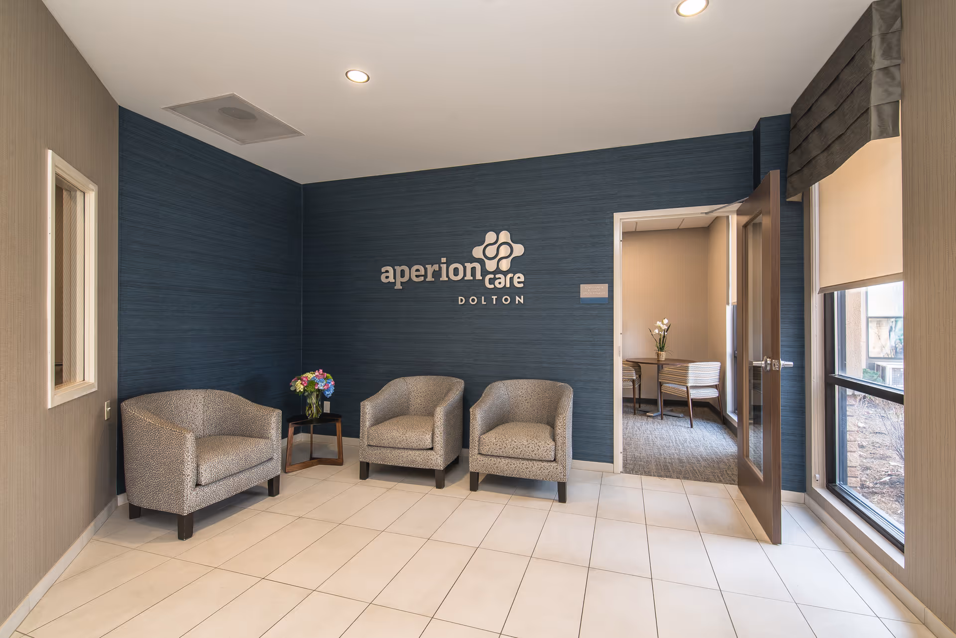 Waiting area inside Aperion Care Dolton facility with three upholstered chairs arranged against a blue accent wall featuring the Aperion Care Dolton logo. A small table with a vase of flowers is placed between two chairs. To the right, an open door leads to another room with a desk and chair, and a large window with a roller shade lets in natural light.