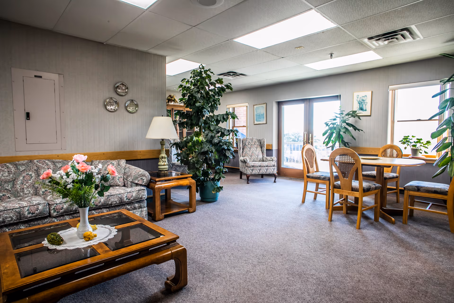 A bright and spacious common area in a senior living facility featuring a floral patterned sofa, a wooden coffee table with a vase of pink flowers, a side table with a lamp, several green plants, a round wooden dining table with four chairs, and large windows and glass doors letting in natural light.