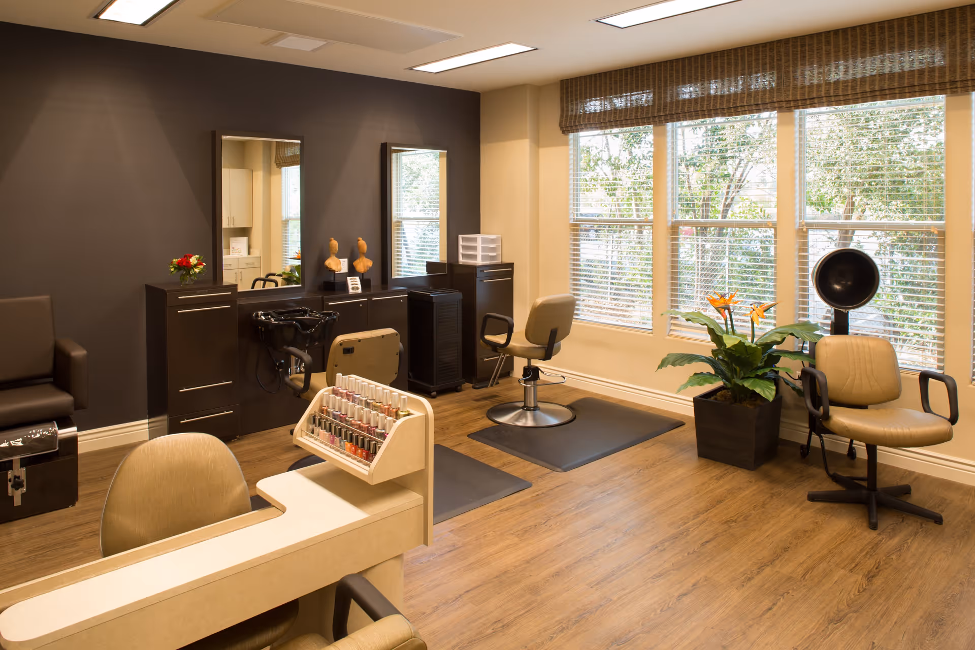 A well-lit salon area inside a senior living facility with two styling chairs in front of mirrors, a hair washing station, a nail polish display, a hair dryer chair, and a large window with blinds letting in natural light. The room has wooden flooring and a potted plant near the window.