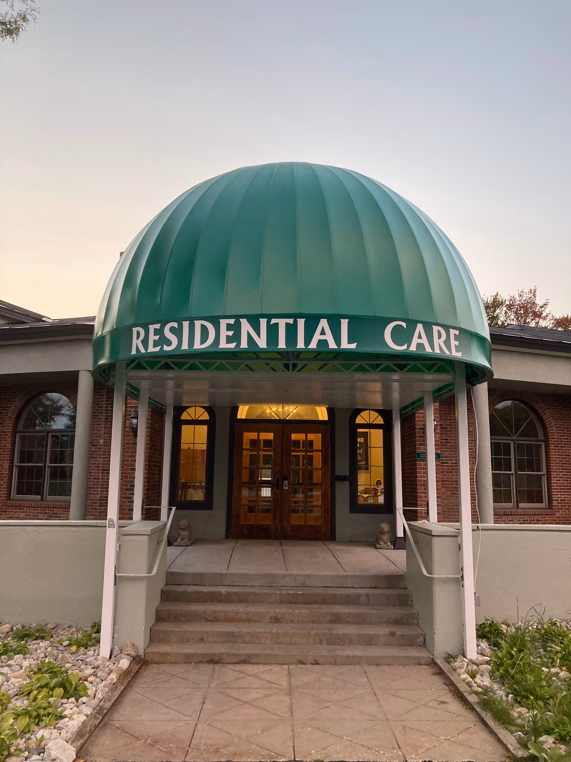 Entrance to a residential care facility with a green dome-shaped awning displaying the words 'RESIDENTIAL CARE'. The entrance has steps leading up to double wooden doors with glass panels, flanked by windows on either side. The building exterior features brick and light-colored walls with some landscaping including rocks and plants.