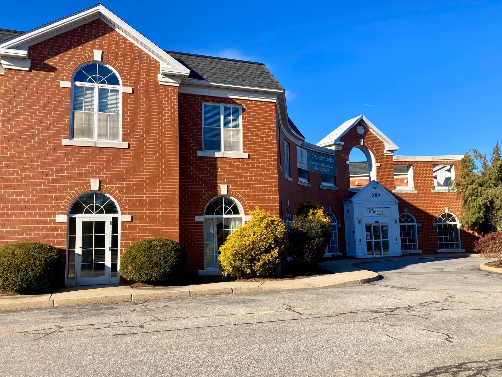 Two-story red brick building with arched windows, shrubs, and a central gabled entrance labeled 589 under a clear blue sky.