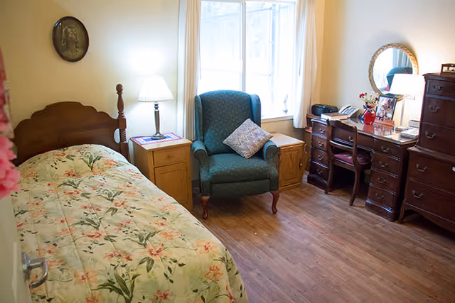 A cozy bedroom with a single bed covered in a floral bedspread, a wooden nightstand with a lamp, a blue upholstered armchair with a decorative pillow, a wooden desk with a chair, a round mirror, and a tall wooden dresser. A large window with white curtains lets in natural light.