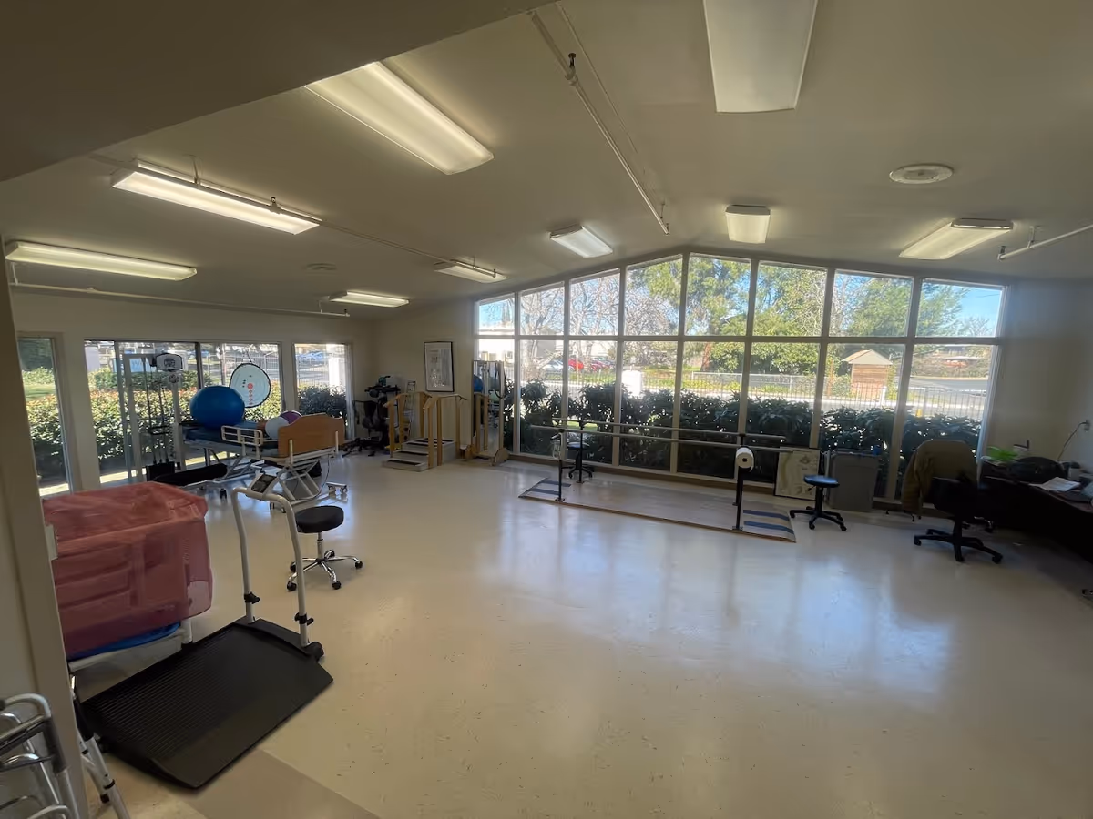 A spacious room with large windows letting in natural light, equipped with physical therapy and rehabilitation equipment including a hospital bed, exercise balls, parallel bars, and various therapy tools. The room has a clean, bright appearance with white floors and ceiling lights.