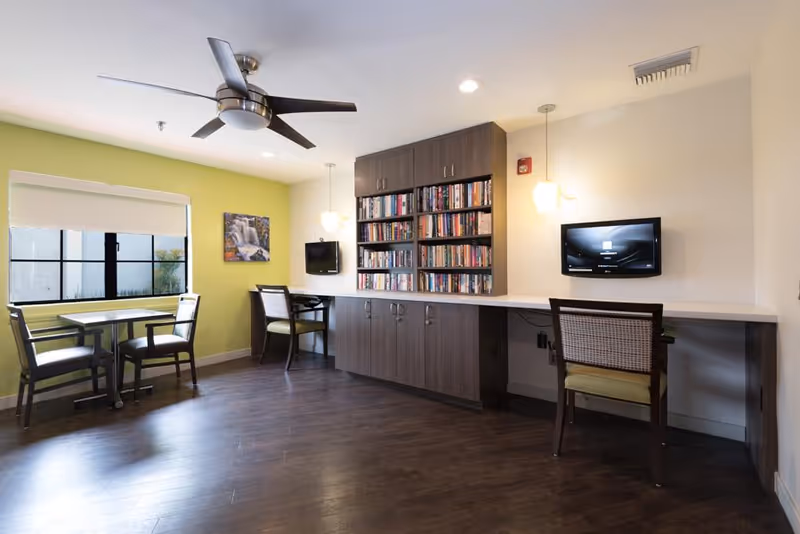 Bright common room with bookshelves, a long counter with chairs and wall-mounted TVs, and a small table by a window.