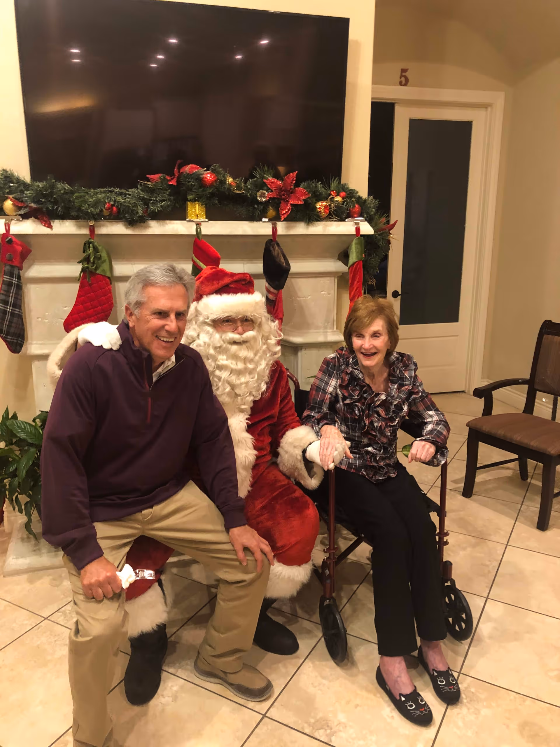 Two older adults sit with a person dressed as Santa Claus in front of a garland-decorated fireplace and large television.