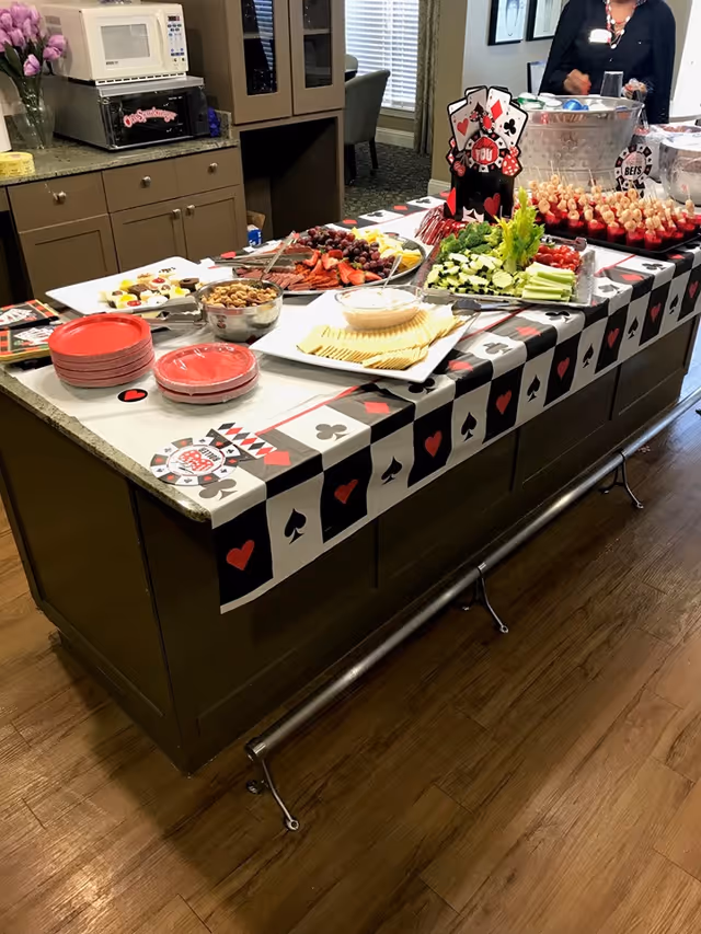 A kitchen island decorated with a playing card themed tablecloth and set up with various snacks including crackers, dip, nuts, grapes, strawberries, celery, cherry tomatoes, and skewers. In the background, there is a microwave, cabinets, and a person standing near a large bowl with ice and drinks.
