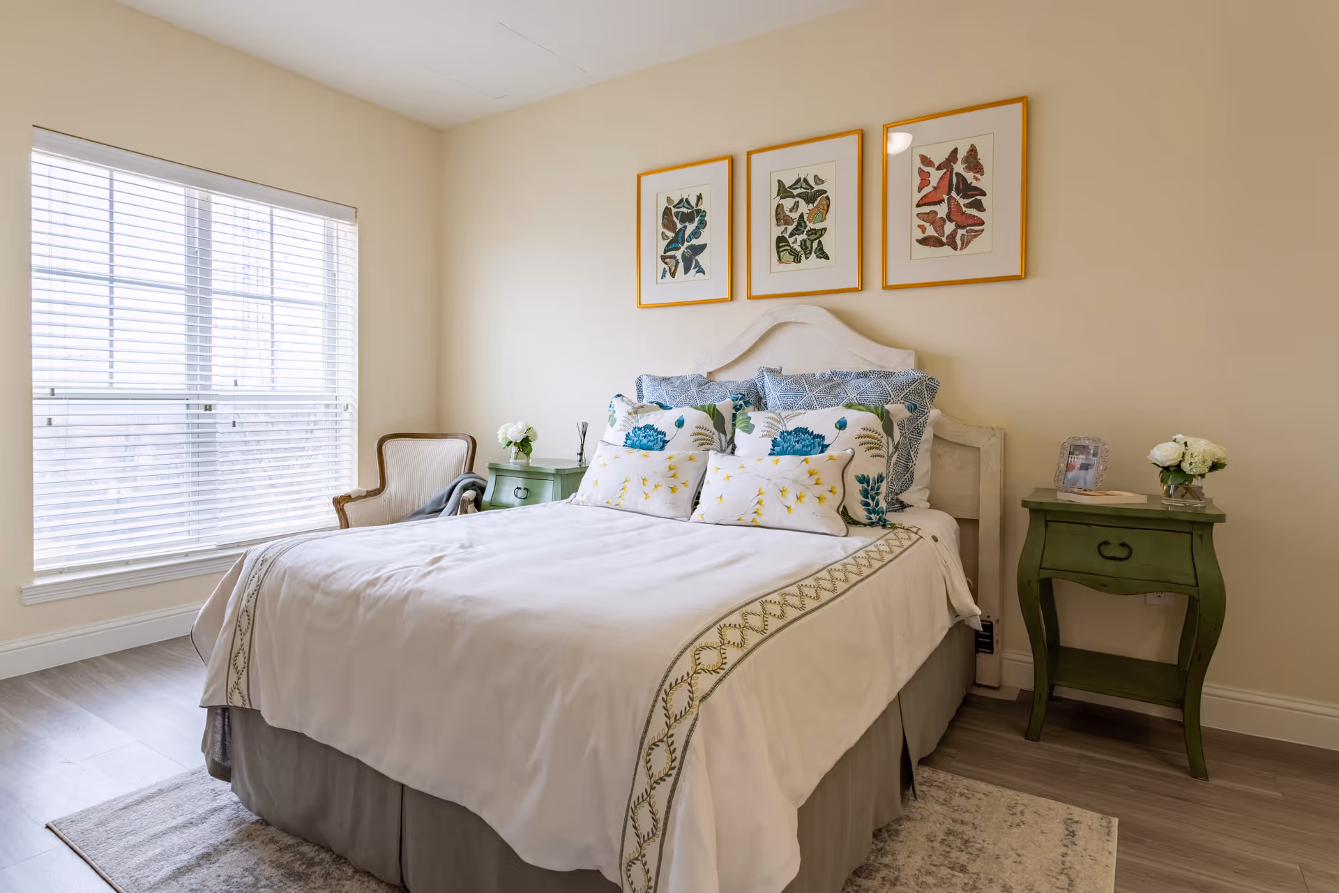 A bright bedroom with a large window covered by white blinds on the left. The bed has a white headboard and is made with white bedding featuring green and blue floral patterns on pillows and a decorative trim on the blanket. There are two green nightstands on either side of the bed, each with a small vase of white flowers. Above the bed are three framed butterfly artworks with gold frames. A beige armchair is positioned near the window, and the floor is covered with light wood flooring and a light-colored rug.