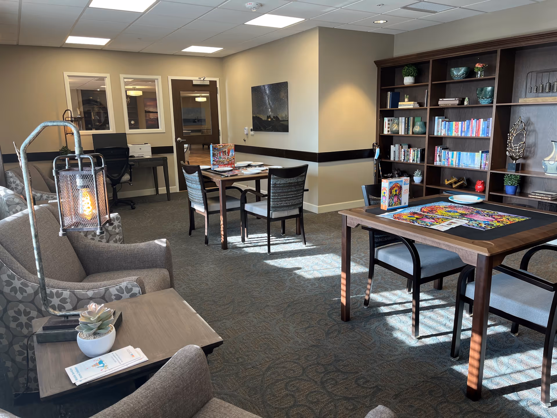 A cozy common room with patterned armchairs, a side table with a small potted succulent and brochures, two tables with chairs, one table holding a colorful puzzle in progress, and a large wooden bookshelf filled with books and decorative items. The room has beige walls, carpeted floor, and ceiling lights.
