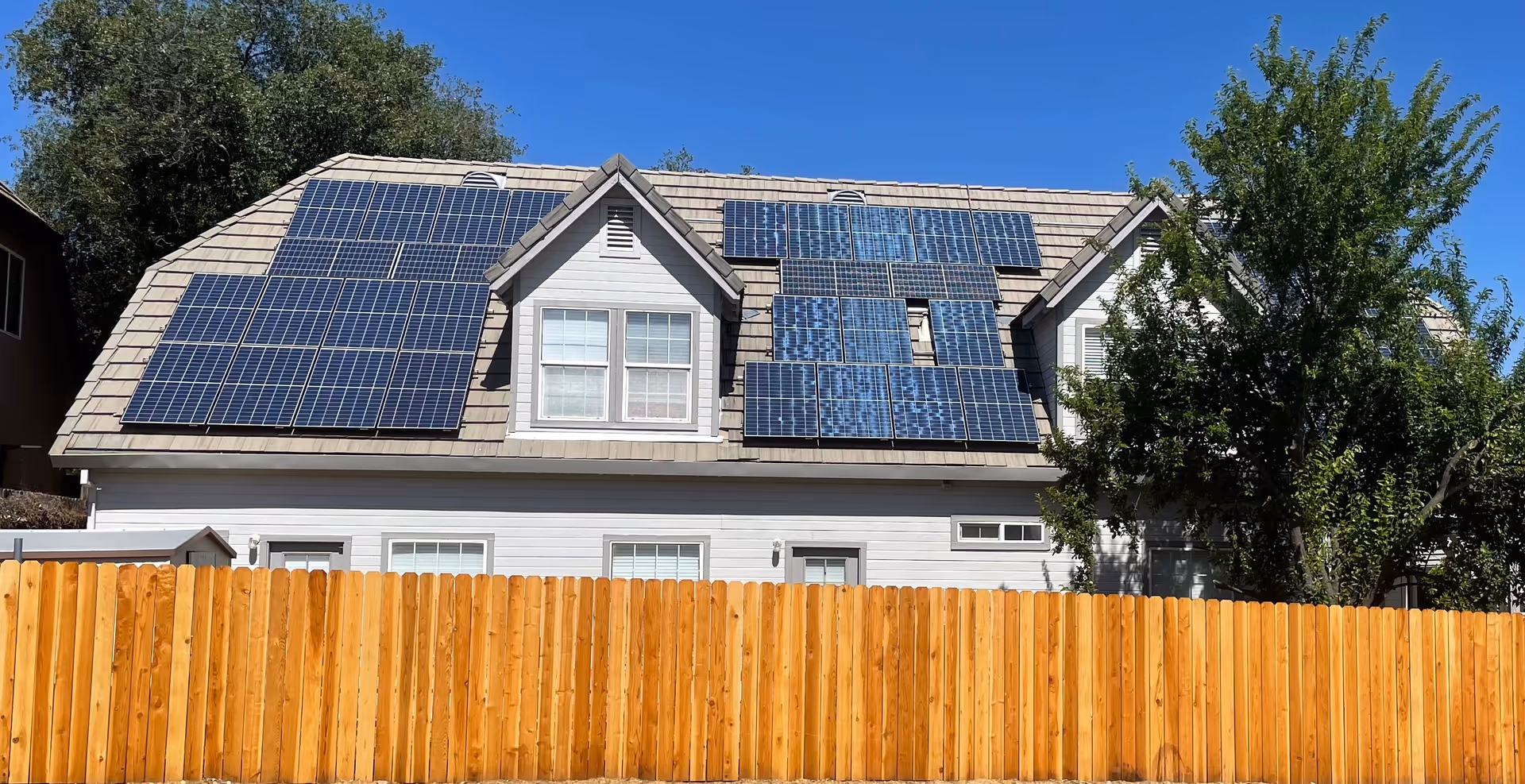 A two-story house with a gray shingled roof covered with multiple solar panels. The house has white siding and several windows. In front of the house is a tall wooden fence, and there are trees on the right side. The sky is clear and blue.