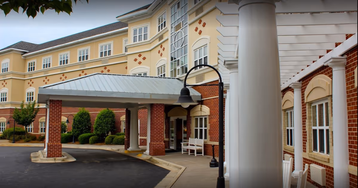 Covered entrance and drive-up portico of a multi-story senior living building with brick columns, white pillars, benches and landscaped grounds.
