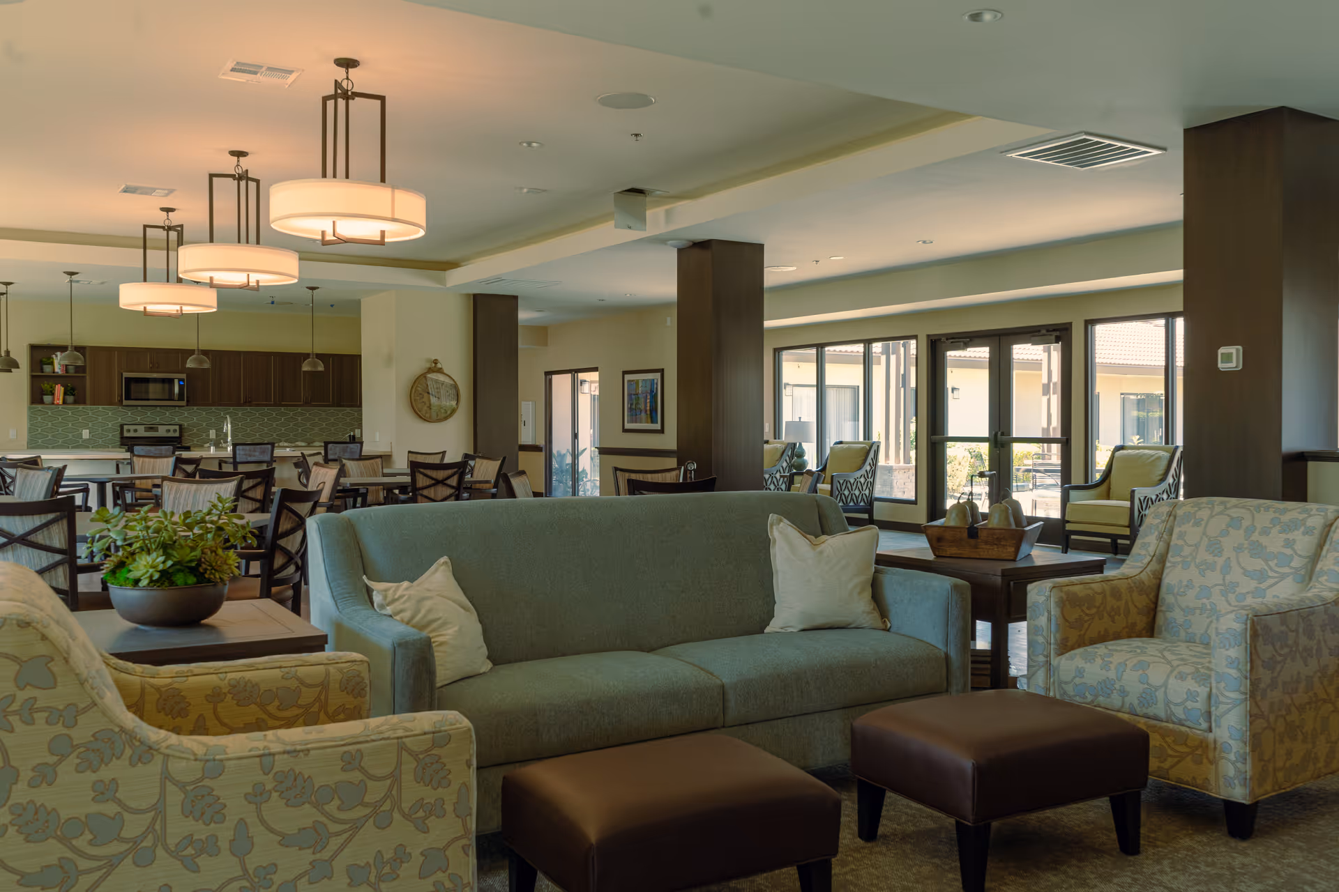 Spacious communal living area with sofas, armchairs, ottomans, and a dining area and kitchen in the background.