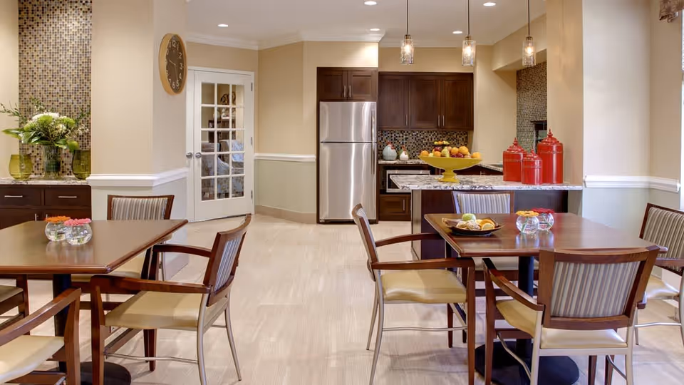 A dining area with wooden tables and chairs featuring beige cushions. In the background, there is a kitchen with dark wood cabinets, a stainless steel refrigerator, a mosaic tile backsplash, and a countertop with decorative items including a yellow fruit bowl and red canisters. The room has light-colored walls and flooring, pendant lights hanging above the kitchen counter, and a clock on the wall near a glass-paneled door.