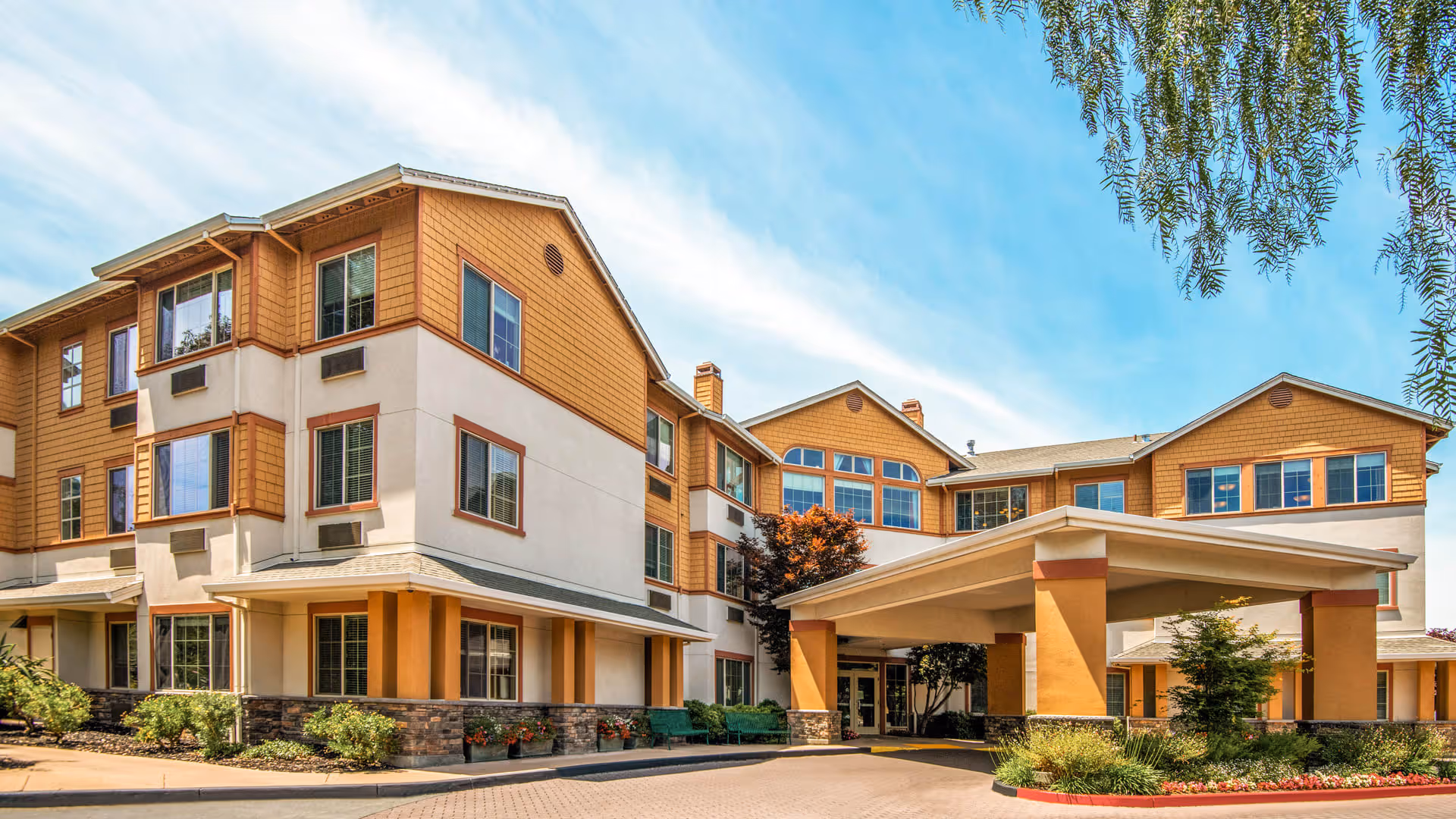 Exterior view of a three-story senior living facility building with a covered entrance, multiple windows, and landscaped greenery including bushes and small trees under a partly cloudy sky.