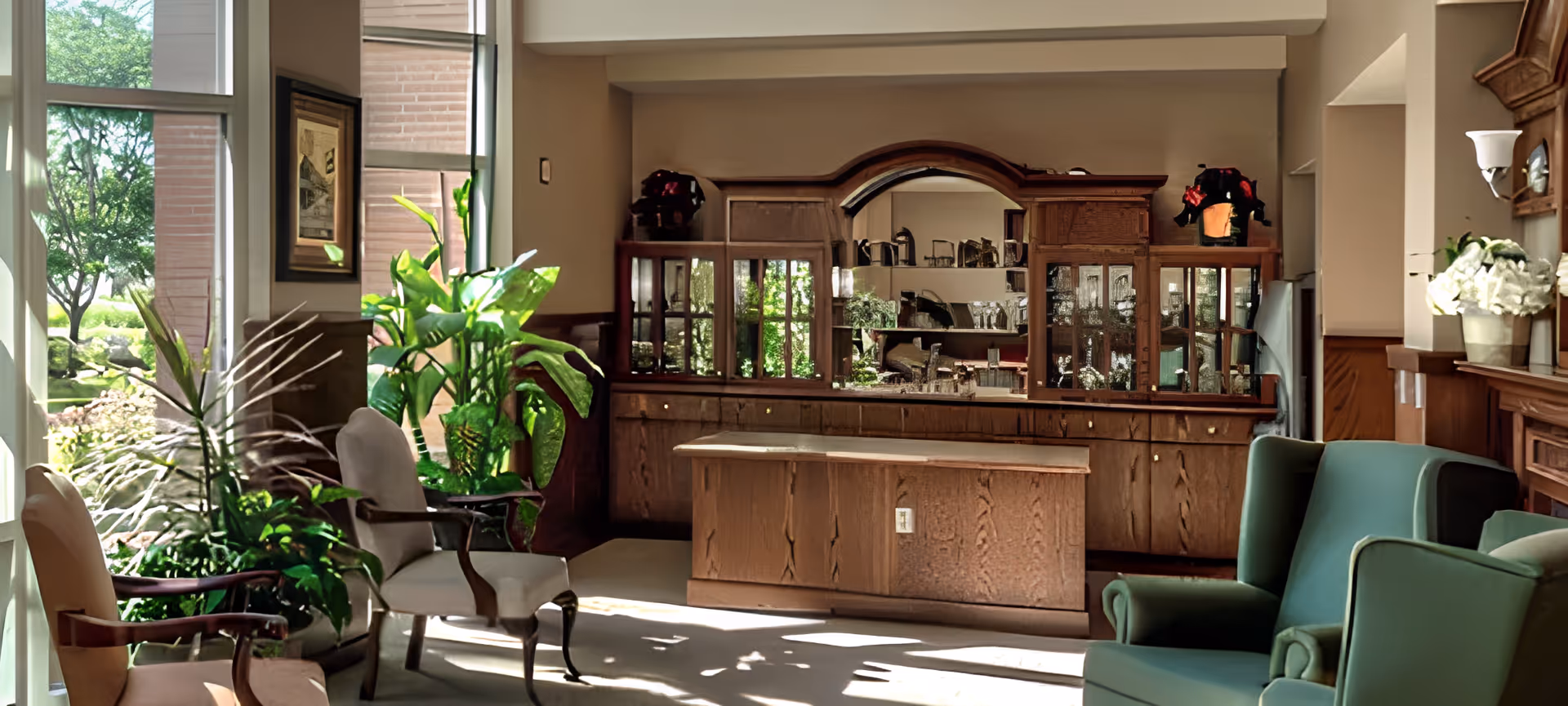 Sunlit common lounge area with upholstered chairs, potted plants, and a wooden cabinet/display behind a central counter.