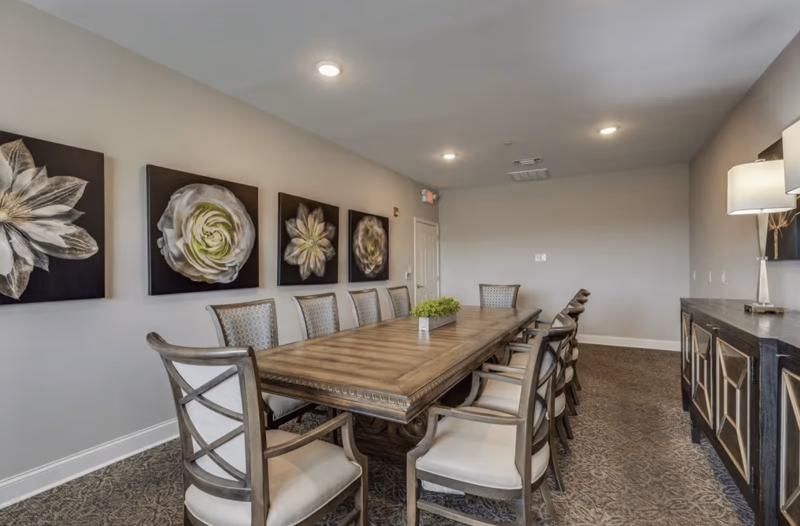 A long wooden dining table with ten cushioned chairs arranged around it in a well-lit room. The walls are decorated with four large floral paintings, and there is a sideboard with a lamp on the right side of the room. The carpet has a patterned design, and the ceiling has recessed lighting.