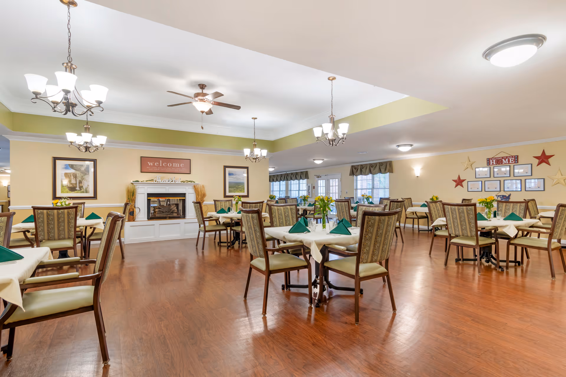 A spacious dining room with multiple tables covered in white tablecloths and green folded napkins. Each table is surrounded by cushioned chairs with patterned upholstery. The room features hardwood flooring, ceiling fans, chandeliers, and a fireplace with a 'welcome' sign above it. The walls are decorated with framed pictures and star-shaped wall hangings.