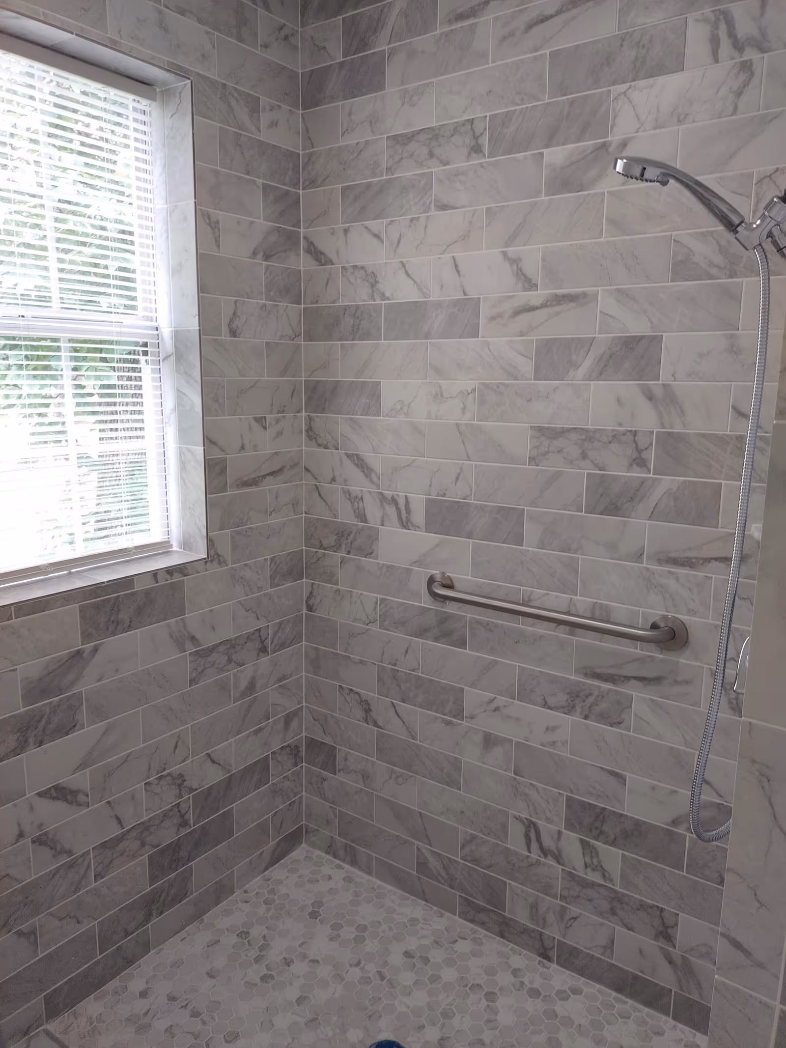 A tiled walk-in shower with gray marble subway tiles, a grab bar, a handheld showerhead, and a window with blinds.