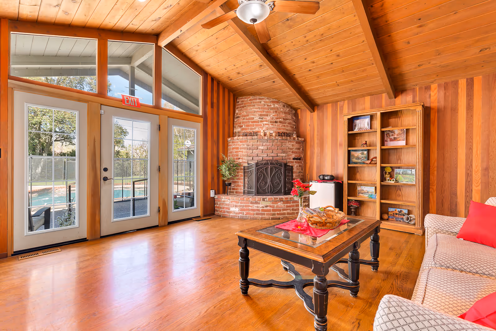 A cozy living room with wooden paneled walls and ceiling, featuring a brick fireplace with a black metal screen. There is a wooden coffee table with a glass top and a red cloth, a basket, and a vase with red flowers. A beige patterned sofa with red cushions is partially visible on the right. A wooden bookshelf with various framed pictures and decorations stands against the wall. Large glass doors and windows let in natural light and provide a view of an outdoor pool area. A ceiling fan with a light fixture hangs from the wooden ceiling.