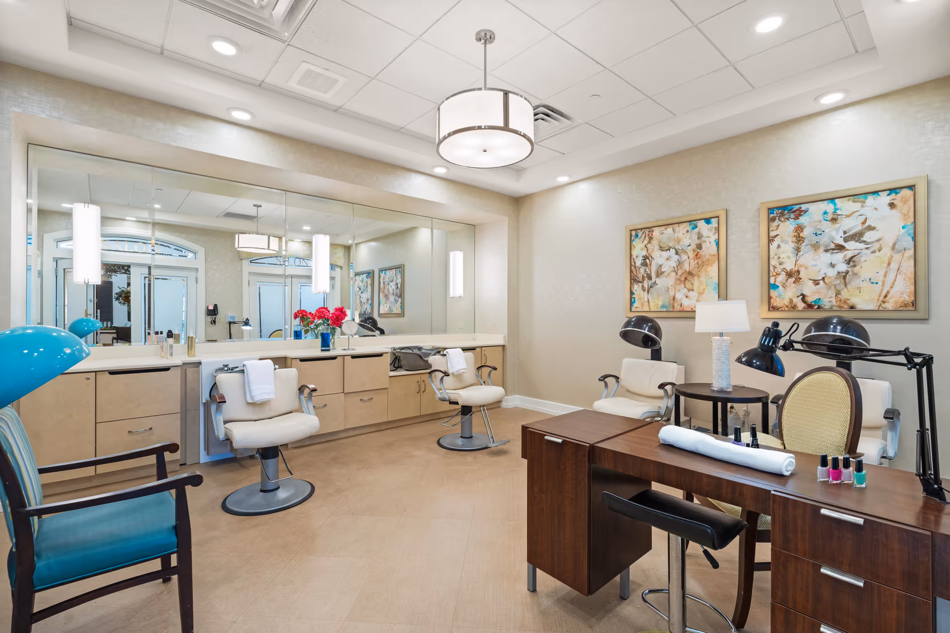 A well-lit salon room in a senior living facility with beige walls and flooring. The room features salon chairs in front of a large mirror, a wooden manicure table with nail polish bottles and a towel, two hair drying chairs with hooded dryers, a blue armchair, and two framed floral paintings on the wall. A modern ceiling light fixture hangs overhead.
