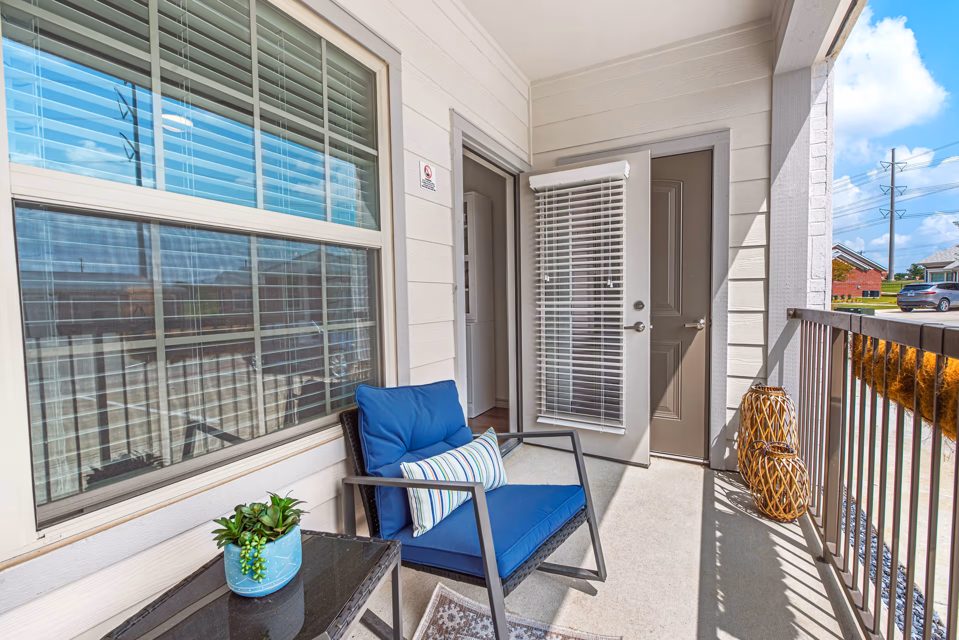 Small apartment balcony with a blue cushioned chair, side table with a potted plant, doors with blinds, and a metal railing.