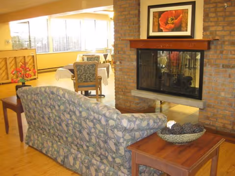 A cozy communal lounge showing a floral-patterned sofa facing a brick fireplace with wooden side tables and dining tables and chairs in the background.