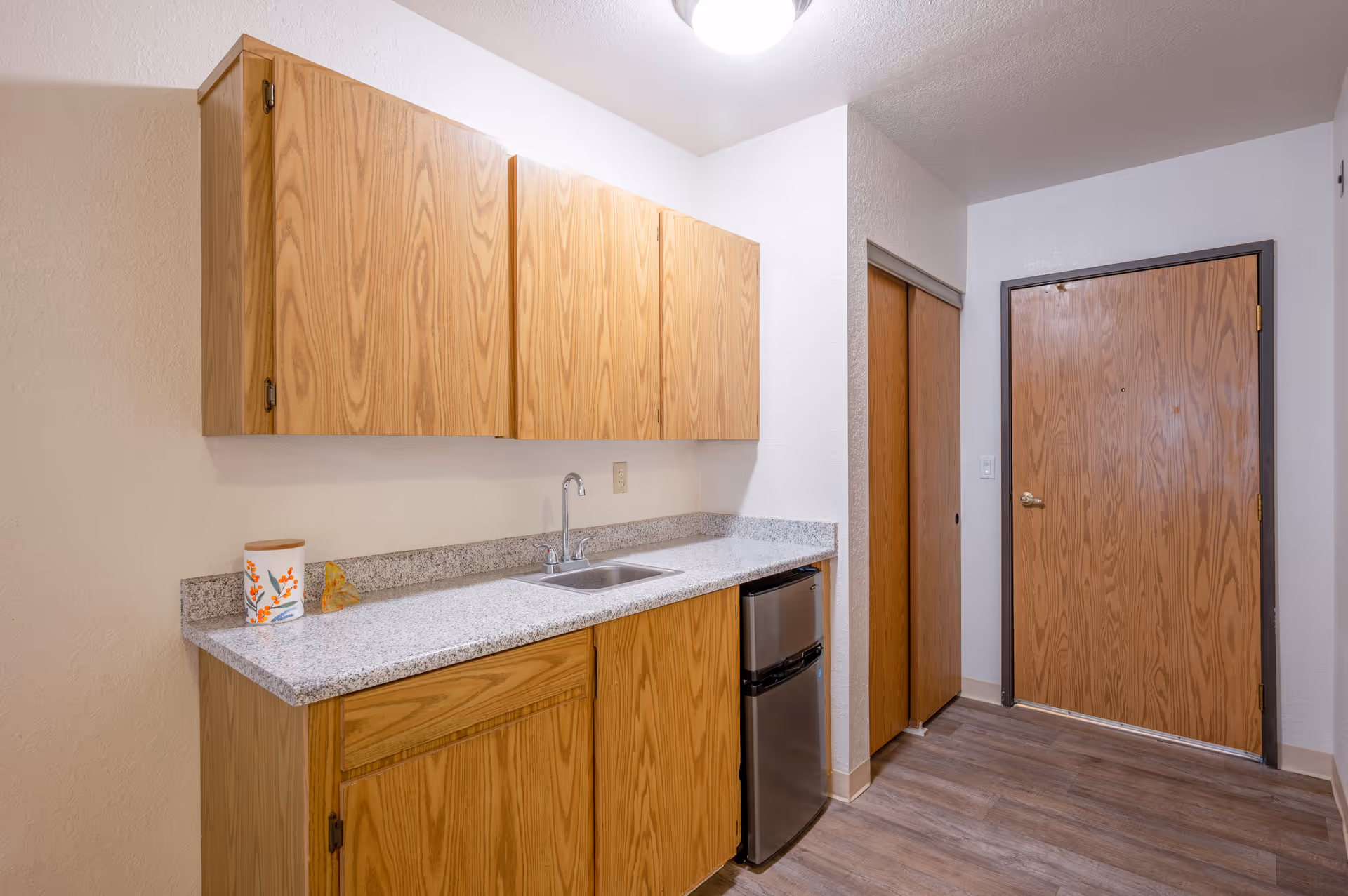 Small kitchenette with wood cabinets, a granite countertop, sink, mini refrigerator and an entry door.