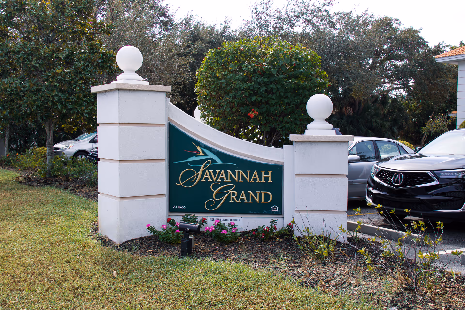 Outdoor image showing a large sign for Savannah Grand, an assisted living facility, set between two white pillars with decorative white spheres on top. The sign is green with gold lettering and a bird graphic. There are cars parked in the background and trees surrounding the area.