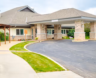 Exterior view of St. Mary's Care Center showing the entrance with a covered drop-off area supported by stone pillars, a curved driveway, and a sidewalk with green grass on the side.