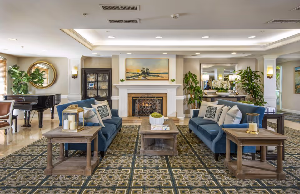 A spacious and elegantly decorated living room area with two blue sofas facing each other, a wooden coffee table in the center, and two side tables with candles. There is a fireplace with a painting above it on the far wall, flanked by plants and a glass cabinet. A grand piano is visible on the left side near a round mirror, and the room is well-lit with ceiling lights and wall sconces.