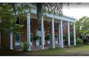 Exterior view of a two-story brick building with large white columns supporting a covered porch. The building is surrounded by trees and greenery, with a grassy area in front.