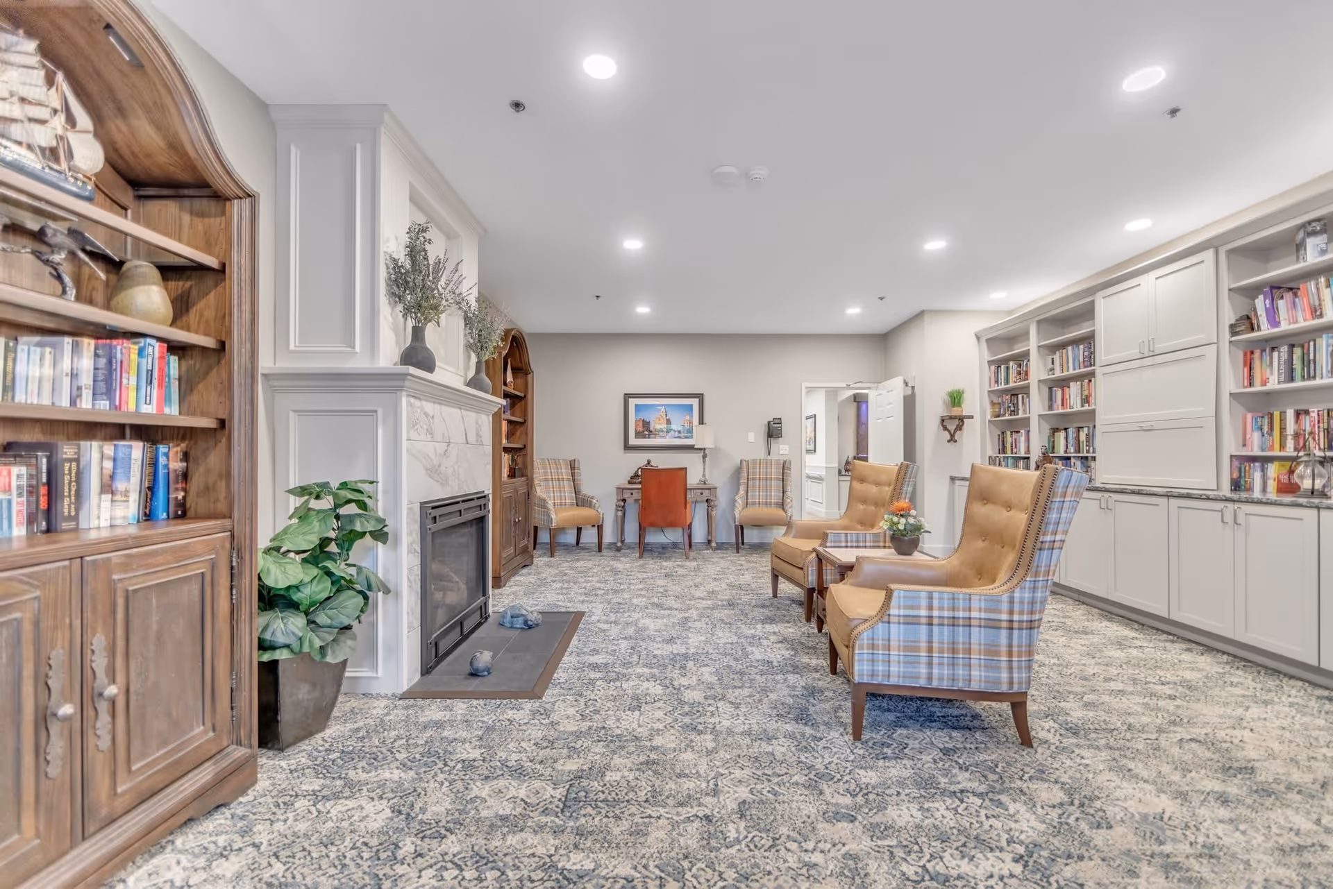 A cozy and well-lit common area in a senior living facility featuring a fireplace with decorative vases on the mantel, wooden bookshelves filled with books, comfortable upholstered chairs with plaid and solid patterns, a small table with a flower arrangement, and a desk with a chair against the far wall. The room has a patterned carpet and recessed ceiling lights.