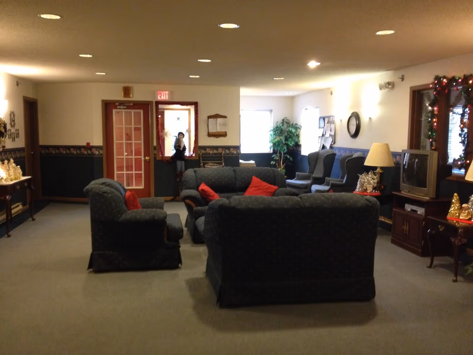 A cozy living room area with dark upholstered sofas and armchairs arranged around a central space. The room has a carpeted floor, a TV on a wooden stand, a lamp, and holiday decorations including garlands and figurines. There are windows letting in natural light and a door with a red frame at the back.