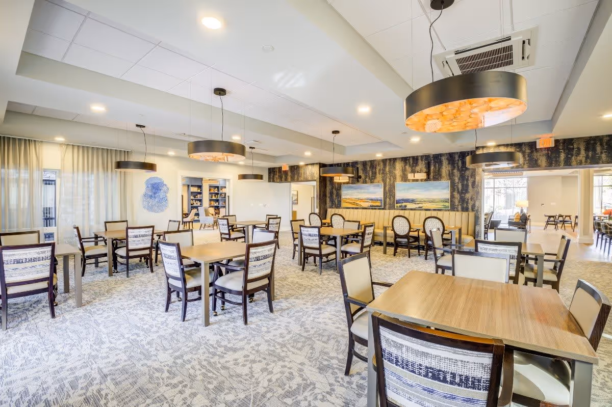 A bright and spacious dining room with multiple wooden tables and chairs arranged neatly. The room features modern pendant lights hanging from the ceiling, patterned carpet flooring, and large windows with sheer curtains allowing natural light to fill the space. There are paintings on the far wall and a built-in bench seating area along one side.