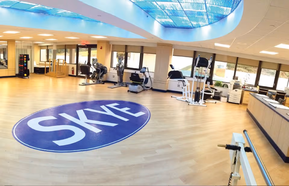 A spacious fitness room with light wood flooring and large windows letting in natural light. The room contains various exercise equipment including stationary bikes, weight machines, and a rack of colorful dumbbells. A large blue circular logo with the word 'SKYE' is displayed prominently on the floor. The ceiling features a skylight allowing additional daylight into the room.