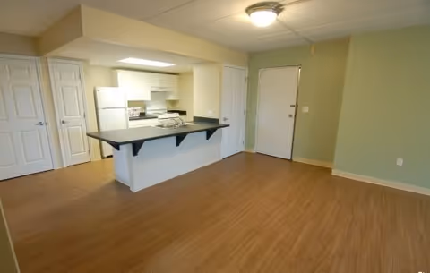 Open living room with wood-look flooring, a kitchen island with a black countertop, white appliances, and an entry door.