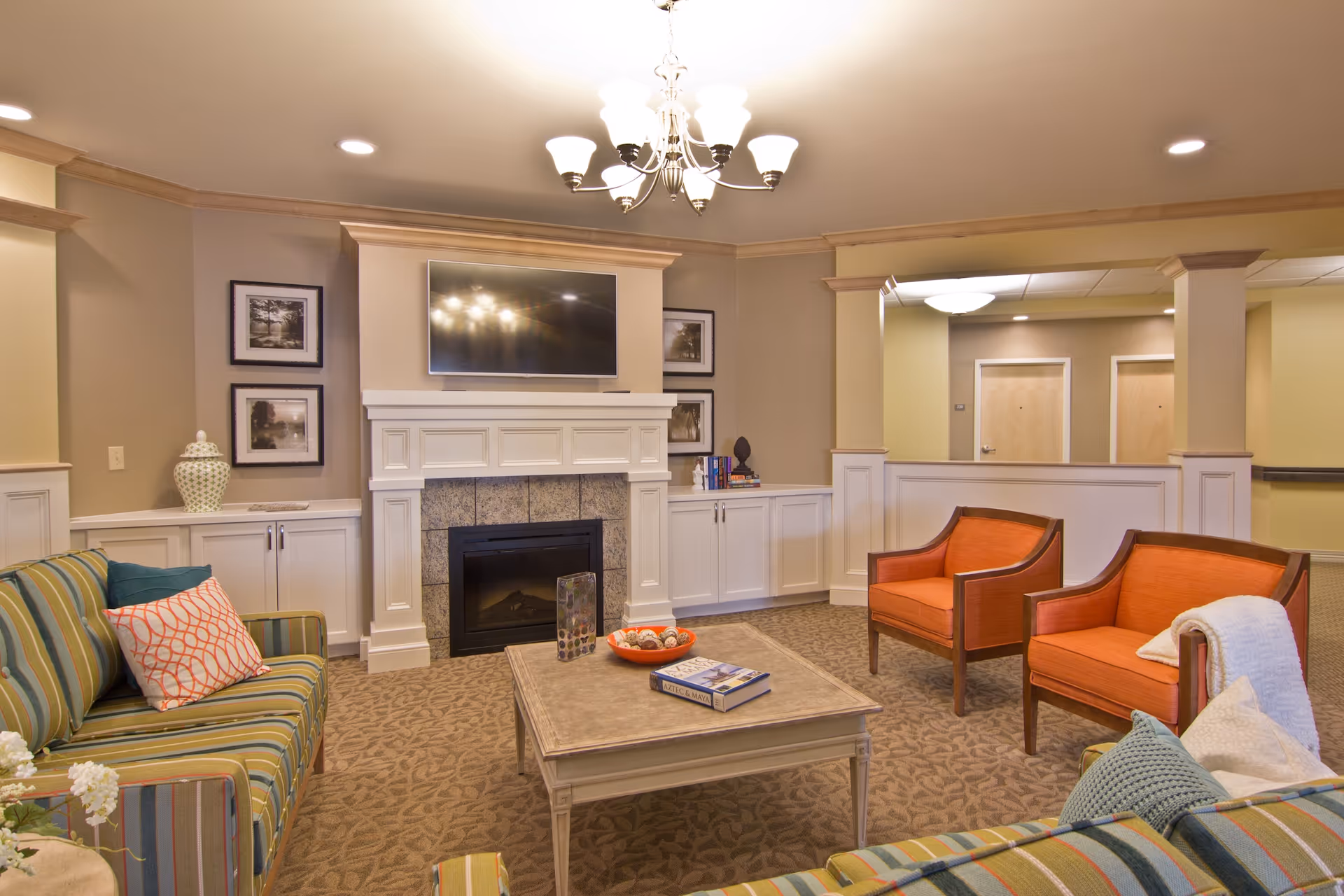 A cozy living room with a striped green sofa, two orange armchairs, a beige coffee table with books and decorative items, a white fireplace with a mounted flat-screen TV above it, and framed pictures on the walls. The room has beige walls, carpeted floor, and a chandelier hanging from the ceiling.