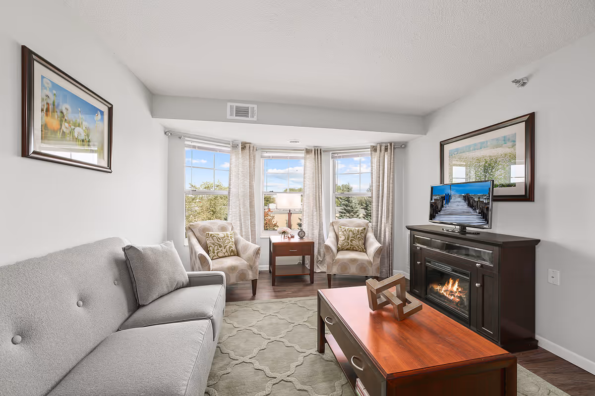 A bright and cozy living room with a gray sofa on the left, two patterned armchairs near three large windows with light curtains, a wooden coffee table with a decorative object, and a dark wooden fireplace with a TV on top displaying an image of a wooden pier leading to the ocean. The walls are light gray with framed artwork hanging on both sides.