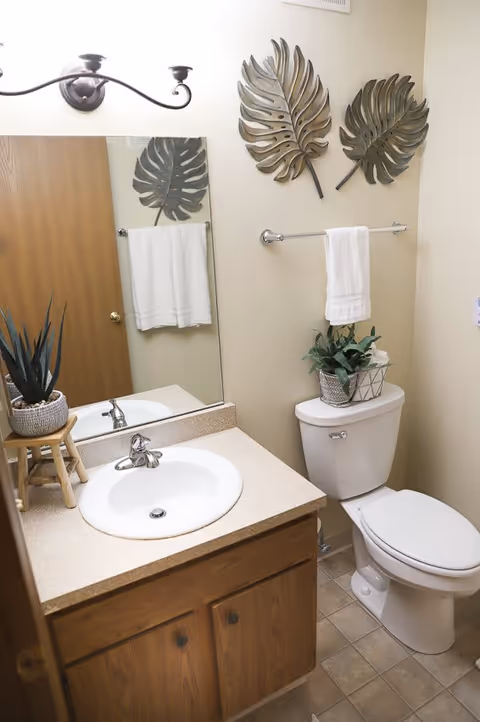 A small bathroom with a white sink set in a beige countertop with wooden cabinets below. Above the sink is a large mirror reflecting a wooden door. On the wall above the toilet are two decorative metal leaf sculptures and a towel rack with a white towel. A potted plant sits on the toilet tank, and another plant is on a small wooden stool next to the sink. The floor is tiled in a light beige pattern.