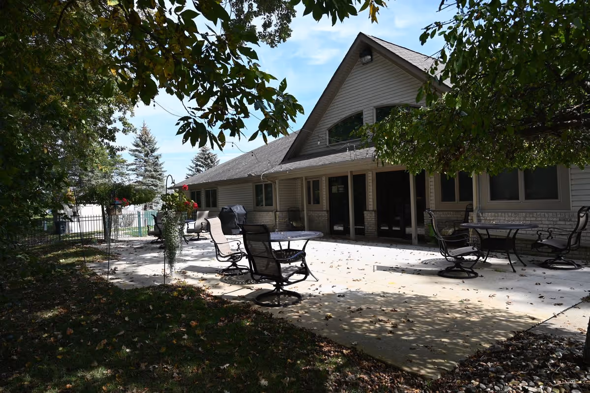 Outdoor patio area at Leisure Manor Residence for Seniors with several metal tables and chairs on a concrete surface, surrounded by trees and greenery, adjacent to a building with large windows and a sloped roof.