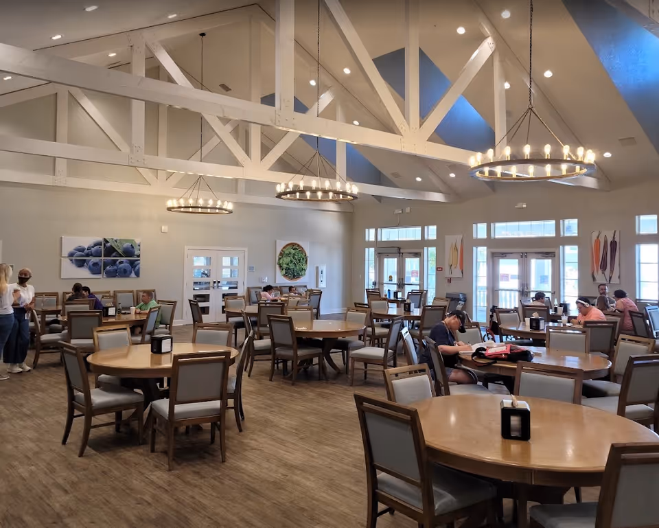 A spacious dining room with multiple round wooden tables and cushioned chairs. Several people are seated at the tables, some engaged in conversation and others reading. The room features high vaulted ceilings with exposed white beams and three large circular chandeliers. Large windows and glass doors allow natural light to fill the space. Colorful fruit and vegetable artwork decorates the walls.